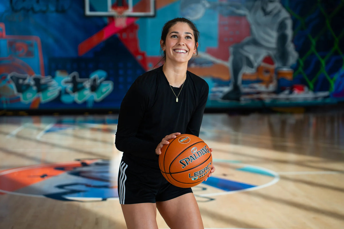 Smiling player holding a Spalding Legacy basketball on a court with a mural